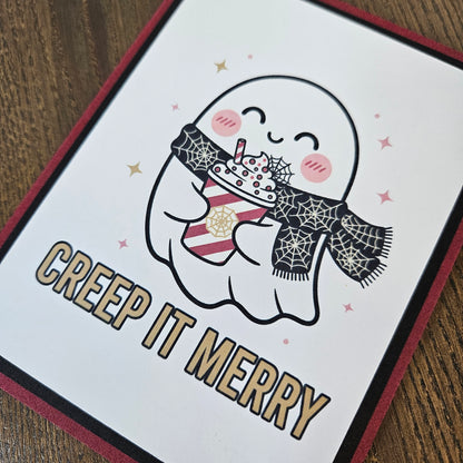 A square greeting card featuring a cute cartoon ghost holding a striped mug with a spiderweb scarf, with the text 'CREEP IT MERRY' in gold outlined letters. The card is photographed at an angle on a wooden surface.