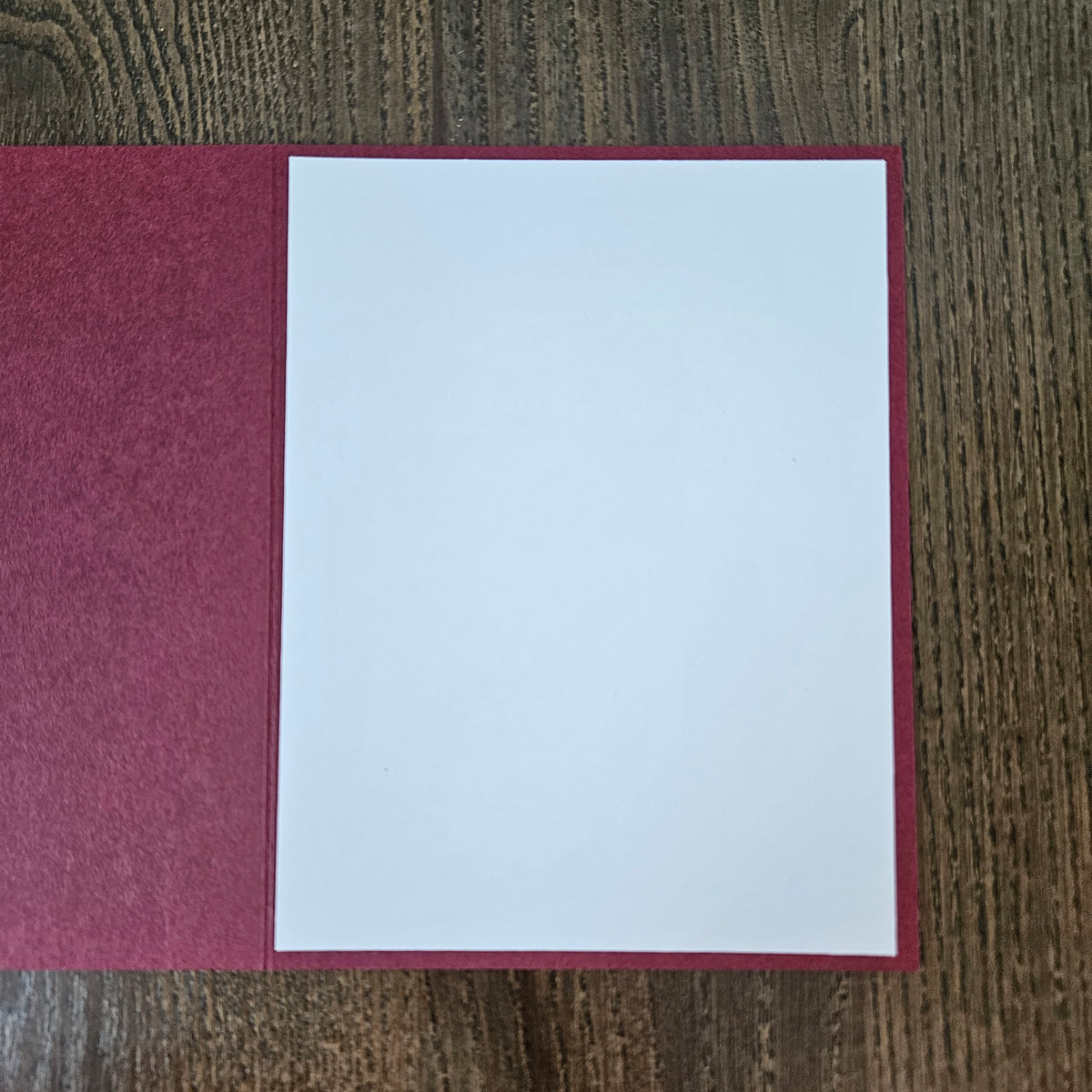A blank white card or letterhead page displayed inside an open burgundy textured folder on a dark wood grain surface.