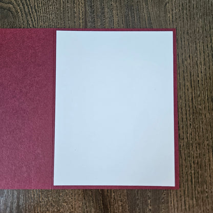 A blank white card or letterhead page displayed inside an open burgundy textured folder on a dark wood grain surface.