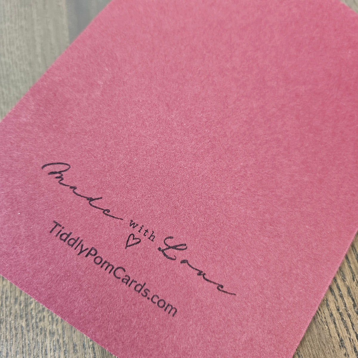 A close-up of a dusty pink textured card with black printed text reading 'Made with Love' and 'TiddlyPomCards.com' on a light wood surface.