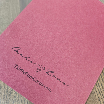A close-up of a dusty pink textured card with black printed text reading 'Made with Love' and 'TiddlyPomCards.com' on a light wood surface.