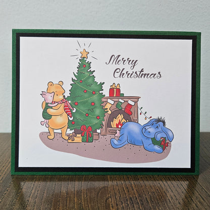 A handmade Christmas greeting card featuring Winnie the Pooh and Tigger by a decorated Christmas tree next to a fireplace with Eeyore resting nearby. The card has a white background with a dark green border and the words 'Merry Christmas' written in black script.
