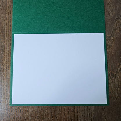 A green felt-covered card holder or envelope with a blank white insert sheet, shown flat on a dark wood surface.