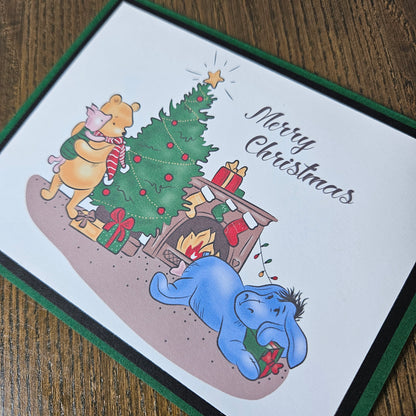 A square Christmas greeting card featuring Winnie the Pooh, Piglet, Tigger, and Eeyore around a decorated tree and fireplace, with the text 'Merry Christmas' on a white cardstock, photographed on a wooden surface.