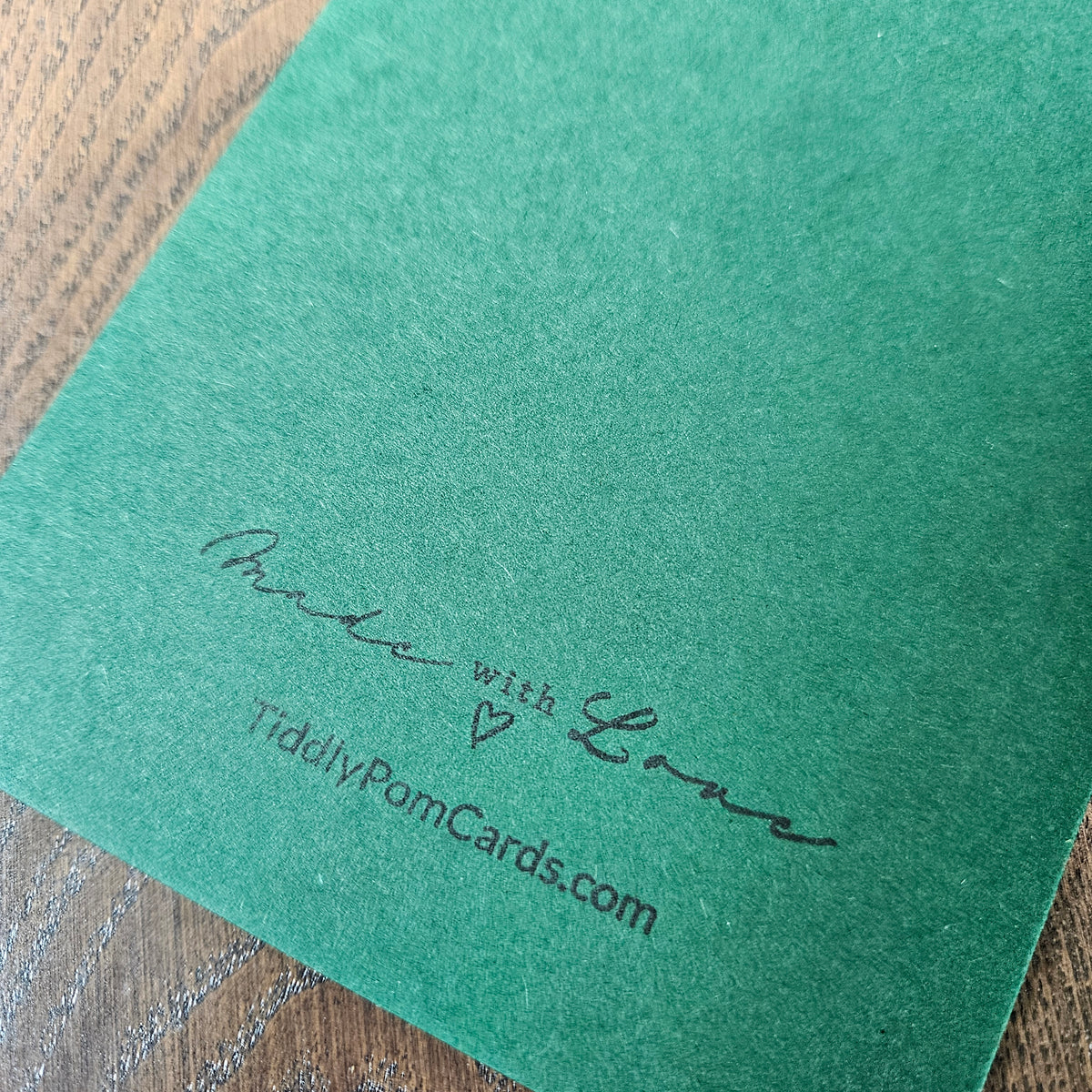 A close-up of a textured green card or envelope corner with the words 'Made with Love' and 'TiddlyPomCards.com' printed in black script, resting on a wooden surface.
