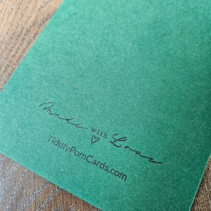 A close-up of a textured green card or envelope corner with the words 'Made with Love' and 'TiddlyPomCards.com' printed in black script, resting on a wooden surface.