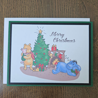 A square Christmas greeting card featuring Winnie the Pooh and Eeyore around a decorated Christmas tree, with 'Merry Christmas' written on the front. The card has a white center, a dark green border, and a black inner border, and is photographed on a brown wooden surface.