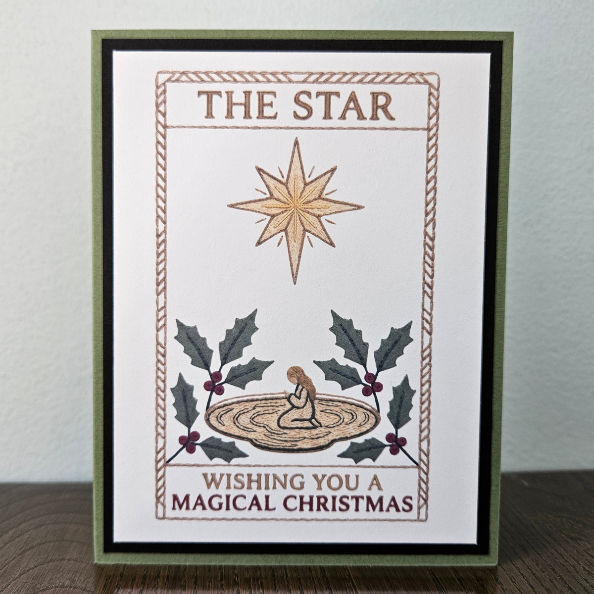 A handmade Christmas greeting card featuring a Star of Bethlehem illustration, holly leaves, and the text 'THE STAR' and 'WISHING YOU A MAGICAL CHRISTMAS', displayed on a wooden surface against a light gray wall.
