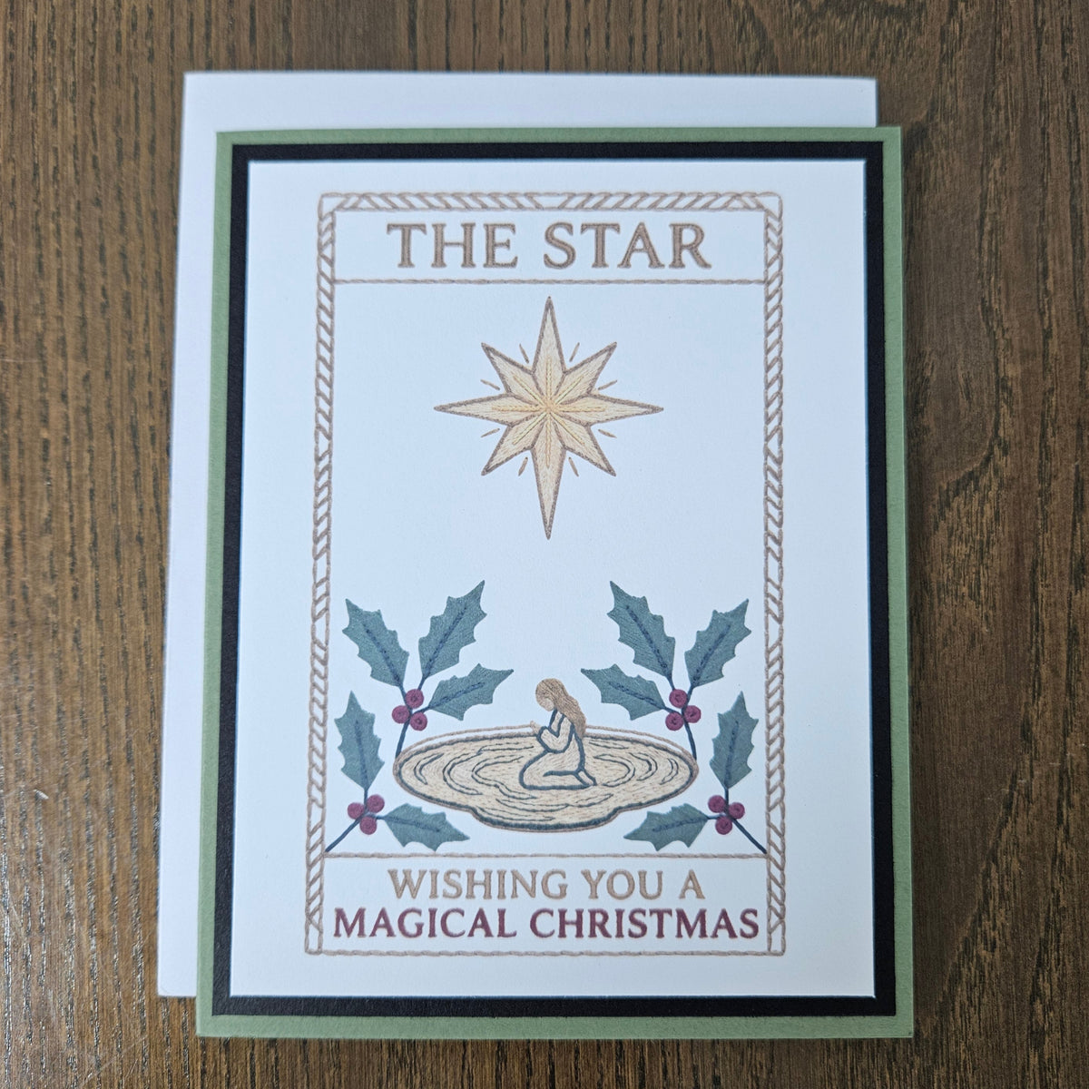 A handmade Christmas greeting card featuring an illustration of 'The Star' with a kneeling baby, holly leaves, and the text 'Wishing You A Magical Christmas', displayed on a wooden table with its white envelope behind it.