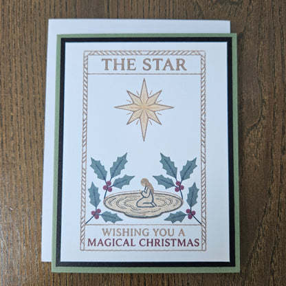 A handmade Christmas greeting card featuring an illustration of 'The Star' with a kneeling baby, holly leaves, and the text 'Wishing You A Magical Christmas', displayed on a wooden table with its white envelope behind it.