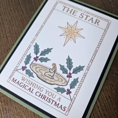 A square Christmas greeting card with a white front, featuring an illustration of a gold star, holly leaves, berries, and a baby in a manger, with the text 'THE STAR' and 'WISHING YOU A MAGICAL CHRISTMAS.' The card is photographed at an angle on a brown wood surface, with a green and black border visible around the card.