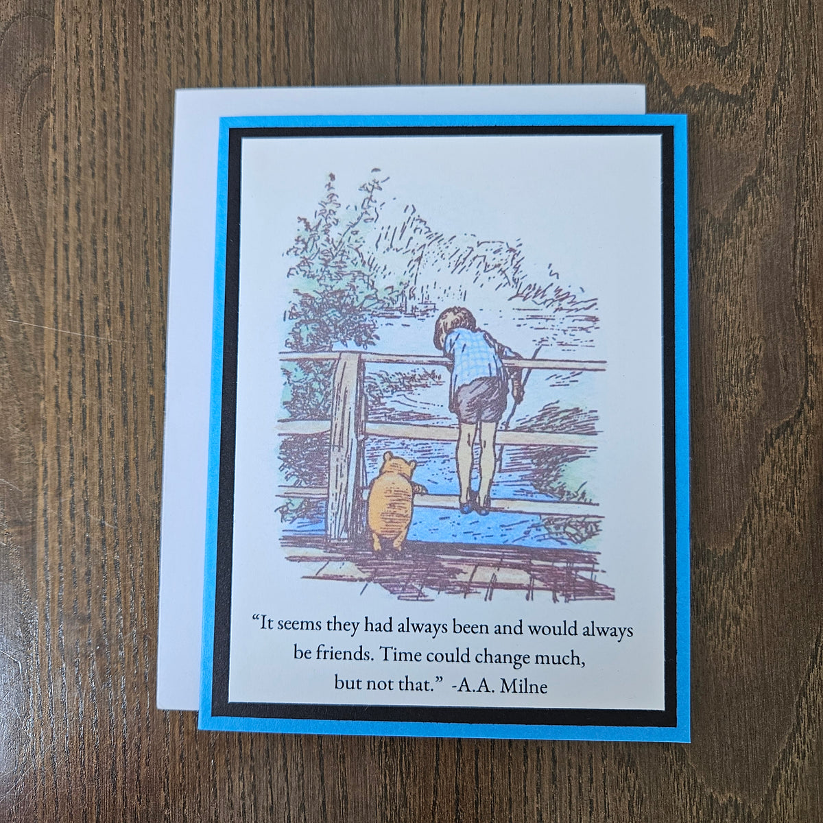 A square greeting card with a white envelope, placed on a brown wooden surface. The card features a blue border and an illustration of Winnie the Pooh and Tigger looking out over water, with the quote, 'It seems they had always been and would always be friends.