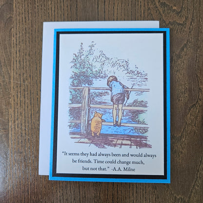 A square greeting card with a white envelope, placed on a brown wooden surface. The card features a blue border and an illustration of Winnie the Pooh and Tigger looking out over water, with the quote, 'It seems they had always been and would always be friends.