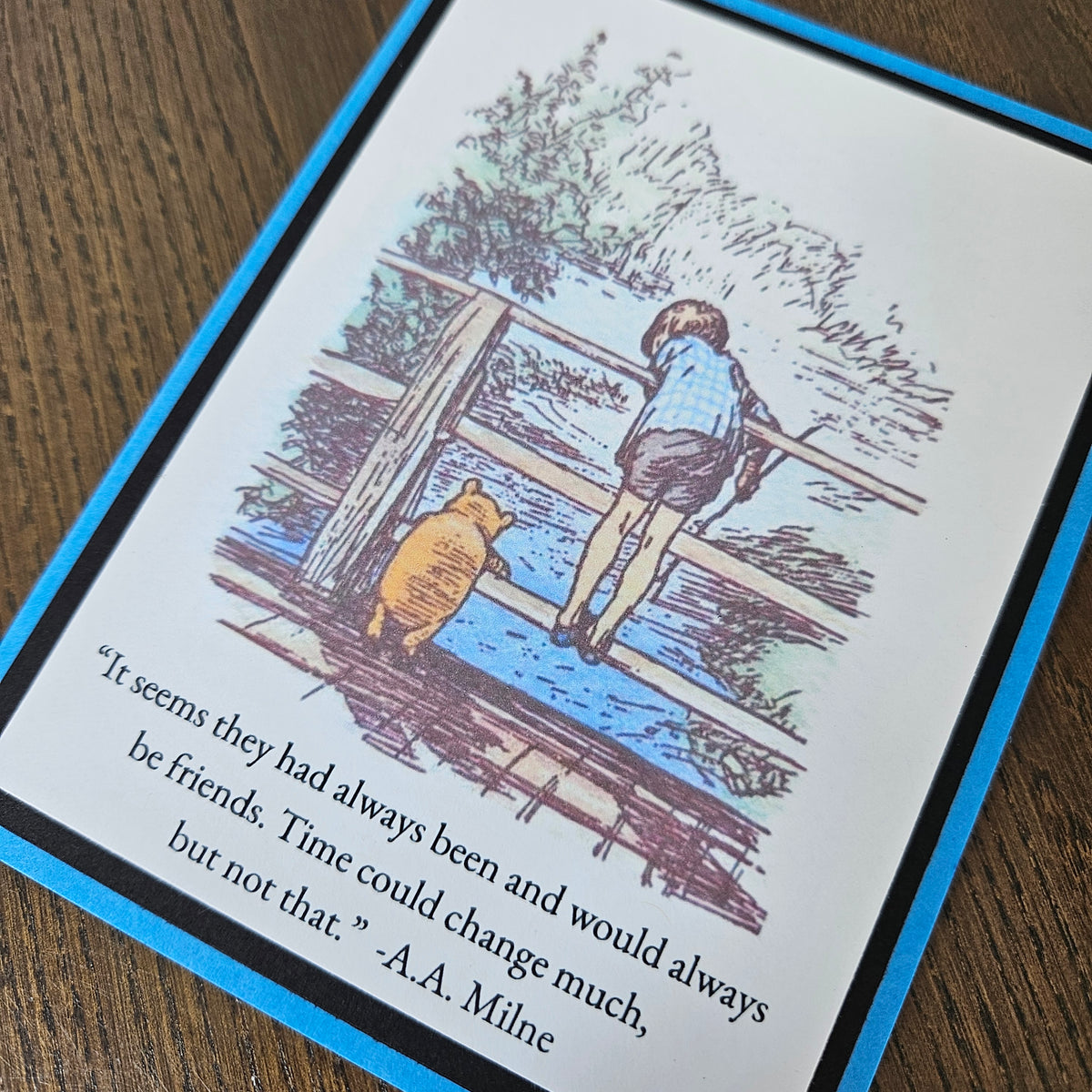 A square printed card or book page featuring a Winnie-the-Pooh illustration with a child and bear by a river, plus a quote from A.A.
