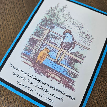 A square printed card or book page featuring a Winnie-the-Pooh illustration with a child and bear by a river, plus a quote from A.A.