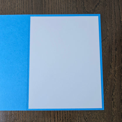 A blank greeting card with a textured bright blue cover and a plain white insert, displayed flat on a dark wood background.