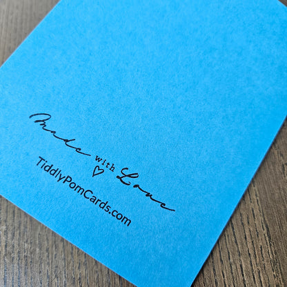 A bright blue square card with black printed text reading 'Made with Love' and 'TiddlyPomCards.com' resting on a brown wood surface.