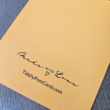 A close-up of a gold-toned greeting card showing the back side with the printed text 'Made with Love' and 'TiddlyPomCards.com' in black script, placed on a blue-gray wood surface.