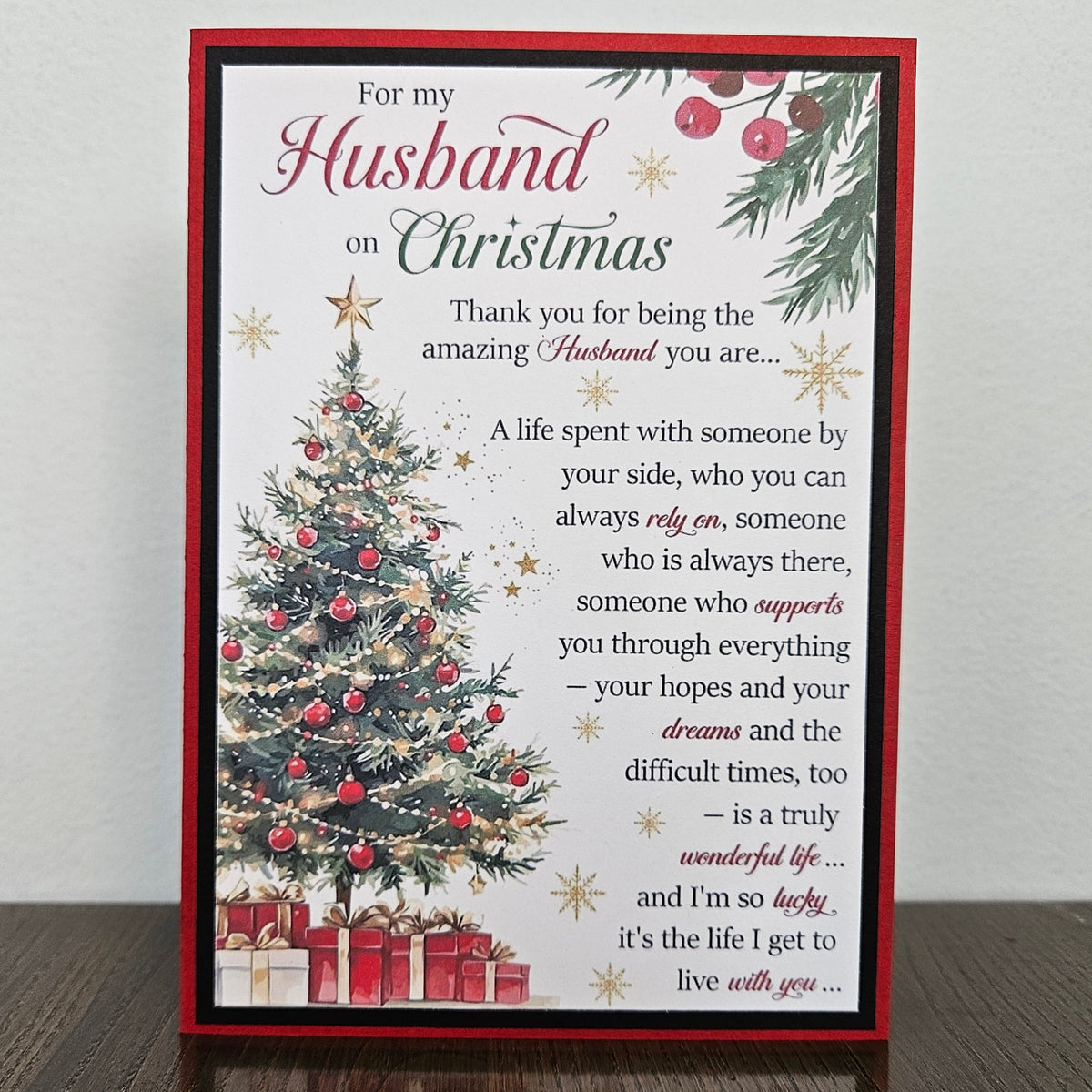 A rectangular Christmas-themed greeting card standing upright on a dark wooden surface against a light wall. The card has a red border, a white center, a decorated Christmas tree, red ornaments, pine branches, and a sentimental message addressed to a husband on Christmas.