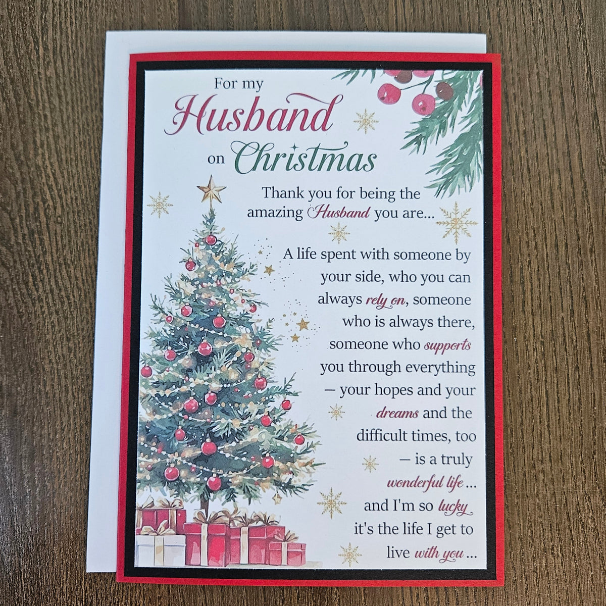 A Christmas-themed husband appreciation card with a red border, featuring a decorated Christmas tree, wrapped gifts, and a sentimental message, displayed on a wooden surface with a white envelope behind it.