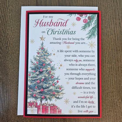 A Christmas-themed husband appreciation card with a red border, featuring a decorated Christmas tree, wrapped gifts, and a sentimental message, displayed on a wooden surface with a white envelope behind it.