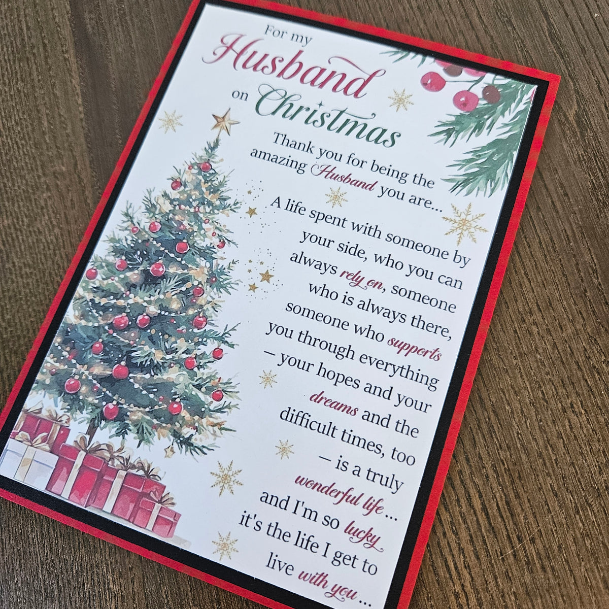 A rectangular Christmas greeting card with a red border, featuring a watercolor Christmas tree, ornaments, gifts, and a sentimental message for a husband, placed on a wooden surface.