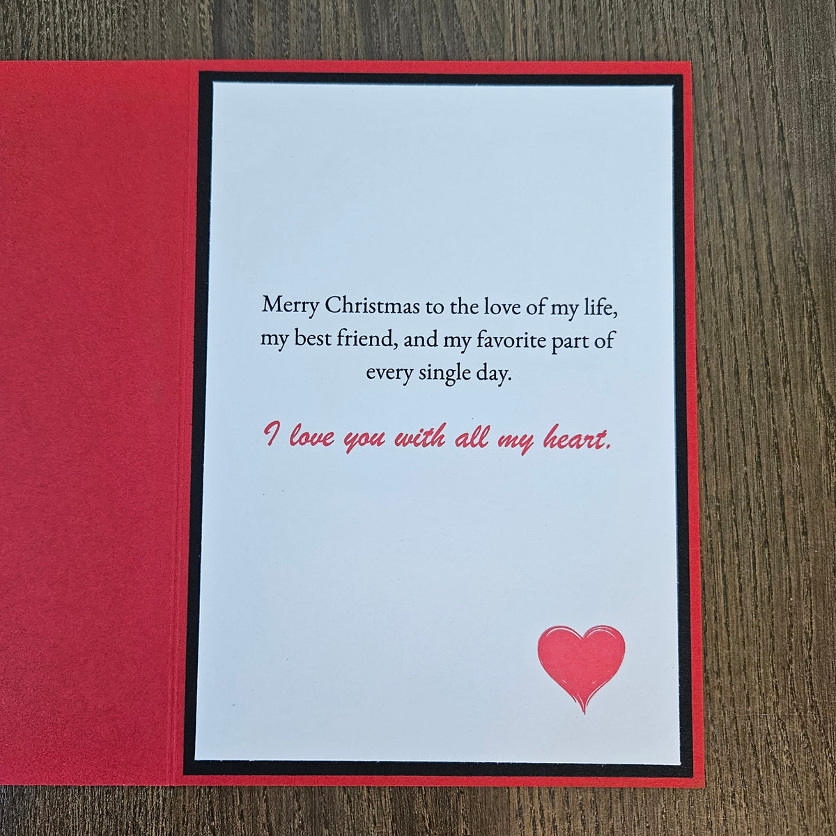 A square Christmas greeting card with a red border and a white center panel, placed on a dark wood surface. The card reads, 'Merry Christmas to the love of my life, my best friend, and my favorite part of every single day.
