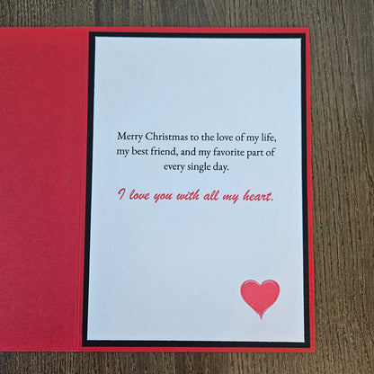 A square Christmas greeting card with a red border and a white center panel, placed on a dark wood surface. The card reads, 'Merry Christmas to the love of my life, my best friend, and my favorite part of every single day.