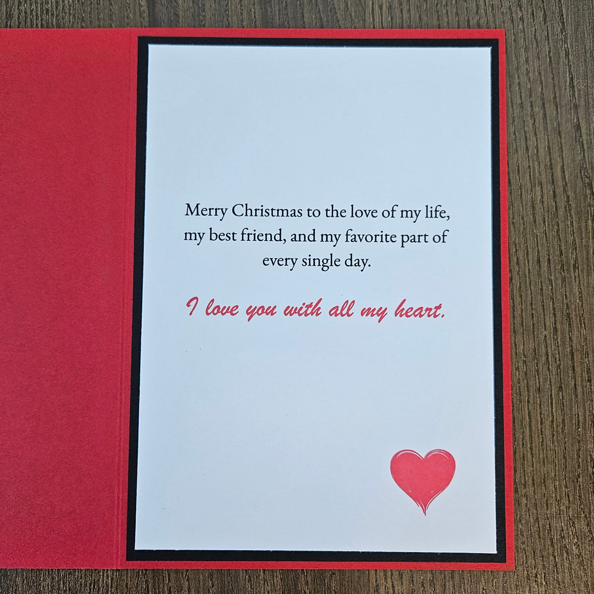A square Christmas greeting card with a red border and a white center panel, placed on a dark wood surface. The card reads, 'Merry Christmas to the love of my life, my best friend, and my favorite part of every single day.