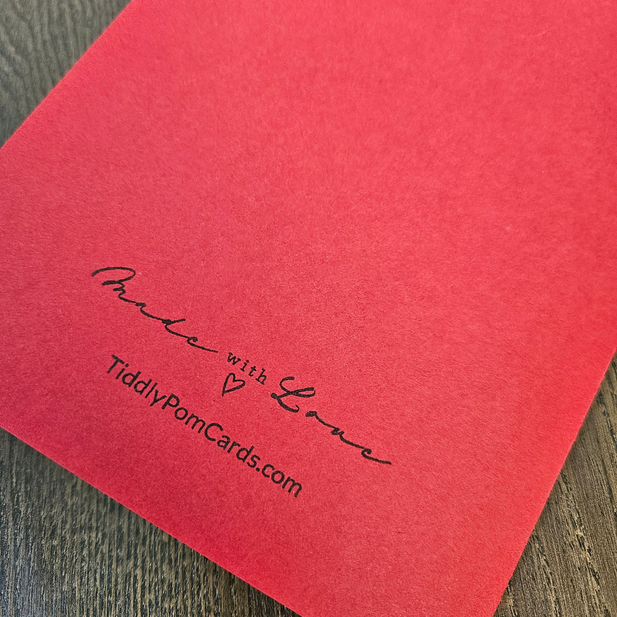 A close-up of a red textured card or paper sheet with black printed text reading 'Made with Love' and 'TidilyPomCards.com' on a dark wood surface.