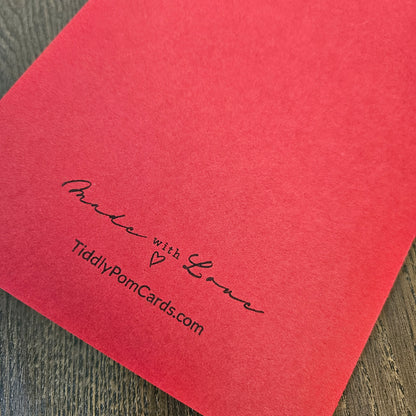 A close-up of a red textured card or paper sheet with black printed text reading 'Made with Love' and 'TidilyPomCards.com' on a dark wood surface.