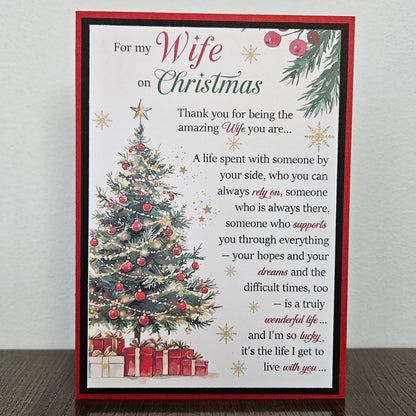 A square Christmas greeting card with a red border, featuring a decorated Christmas tree, wrapped gifts, and a sentimental message for a wife, displayed on a dark wooden surface against a light gray wall.