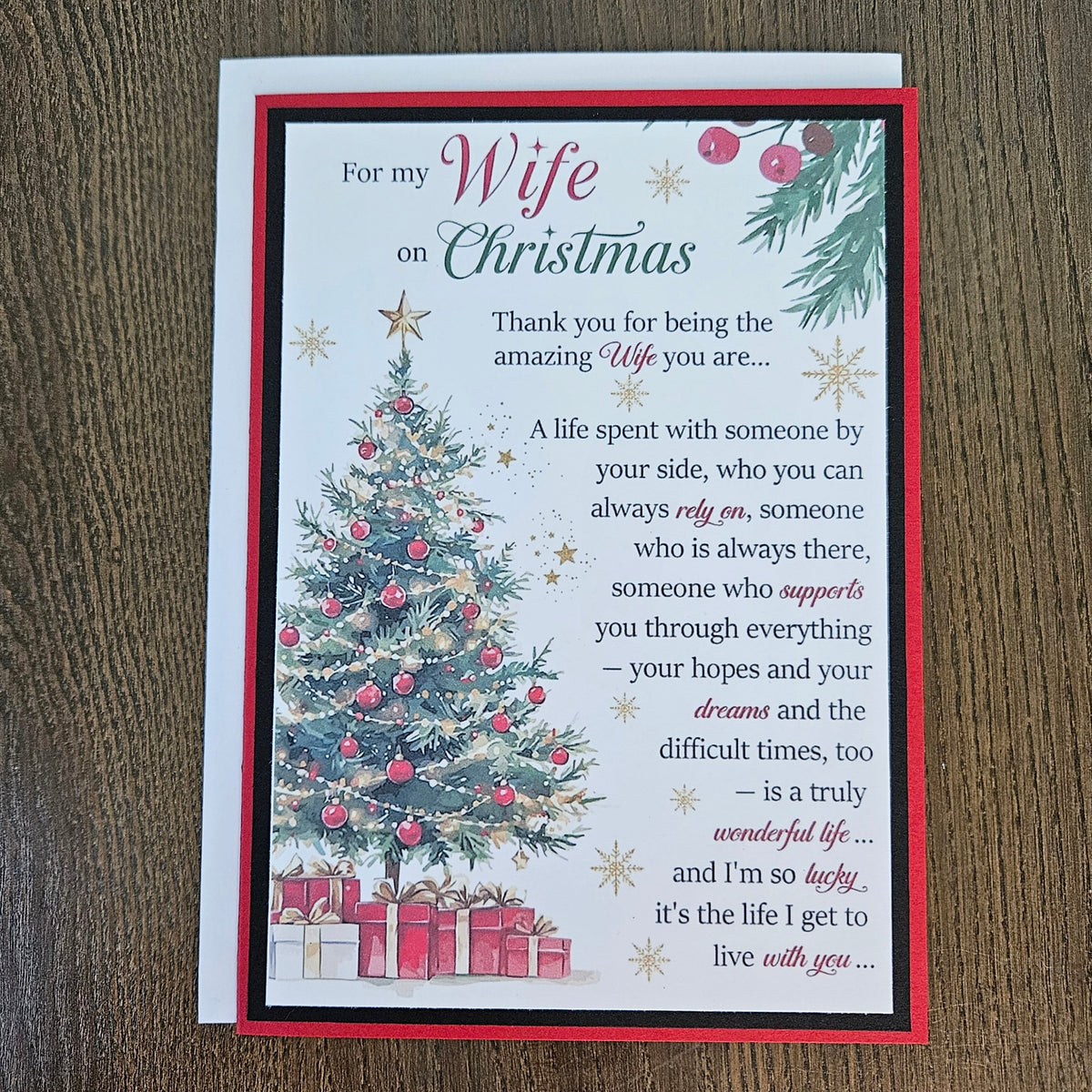A Christmas-themed greeting card for a wife, featuring a white background with a red border, a decorated Christmas tree, wrapped gifts, and a sentimental poem, displayed on a dark wood surface.