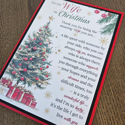 A Christmas-themed 'Wife on Christmas' thank you card with a red border, featuring an illustration of a decorated Christmas tree, wrapped gifts, and a sentimental message, displayed on a wooden surface.