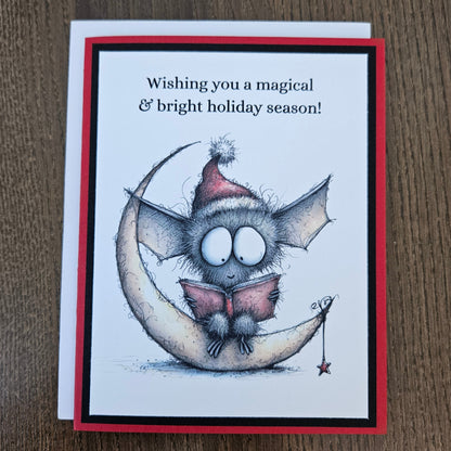 A square greeting card featuring a whimsical illustration of a gray bat wearing a red Santa hat and reading a pink book on a crescent moon, with the text 'Wishing you a magical & bright holiday season!' on a white card with a red and black border, placed on a brown wood surface.