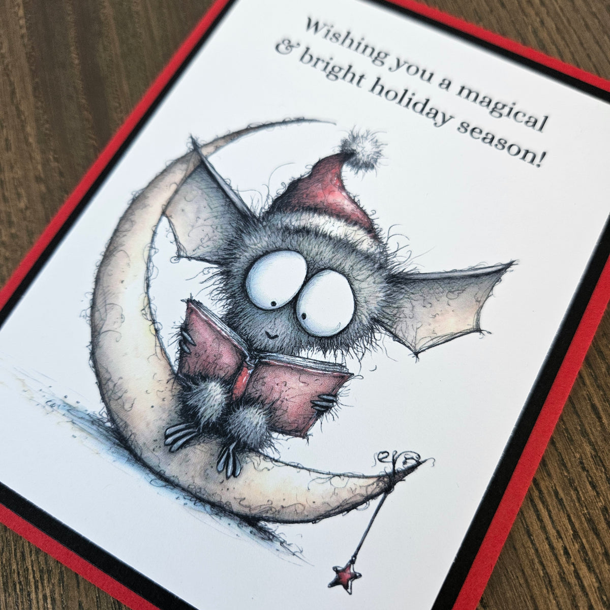 A handmade holiday greeting card featuring a cute bat character sitting on a crescent moon and reading a book, with the text 'Wishing you a magical & bright holiday season!' on a white card with a red border, placed on a wooden surface.
