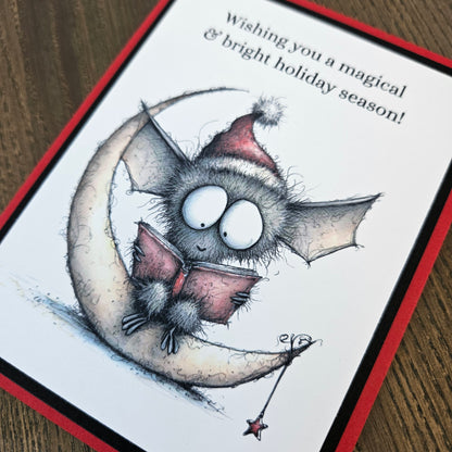 A handmade holiday greeting card featuring a cute bat character sitting on a crescent moon and reading a book, with the text 'Wishing you a magical & bright holiday season!' on a white card with a red border, placed on a wooden surface.