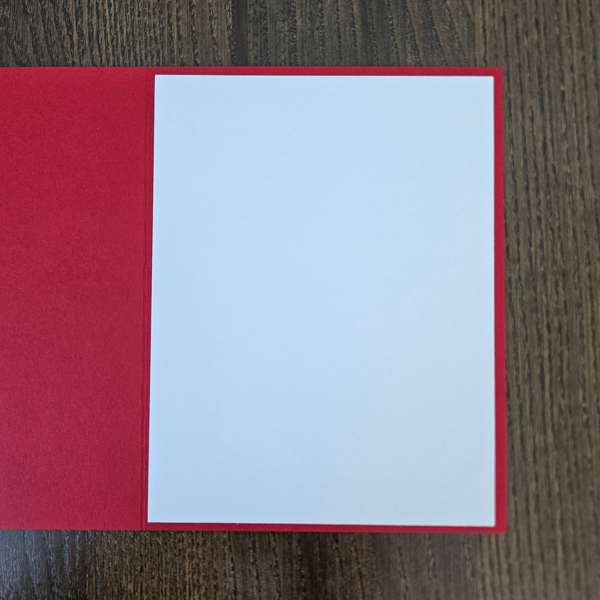 A blank white rectangular card or paper sheet placed on top of a red backing or folder, photographed from above on a dark wood surface.