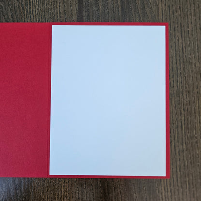 A blank white rectangular card or paper sheet placed on top of a red backing or folder, photographed from above on a dark wood surface.