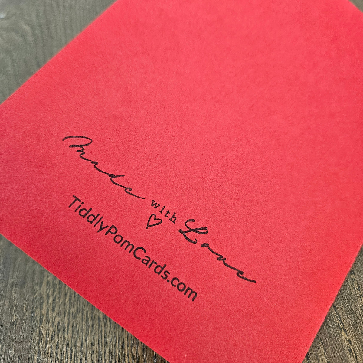 A close-up of a red textured paper card printed with the words 'Made with Love' and the website 'TiddlyPomCards.com' in black script, resting on a dark wood surface.