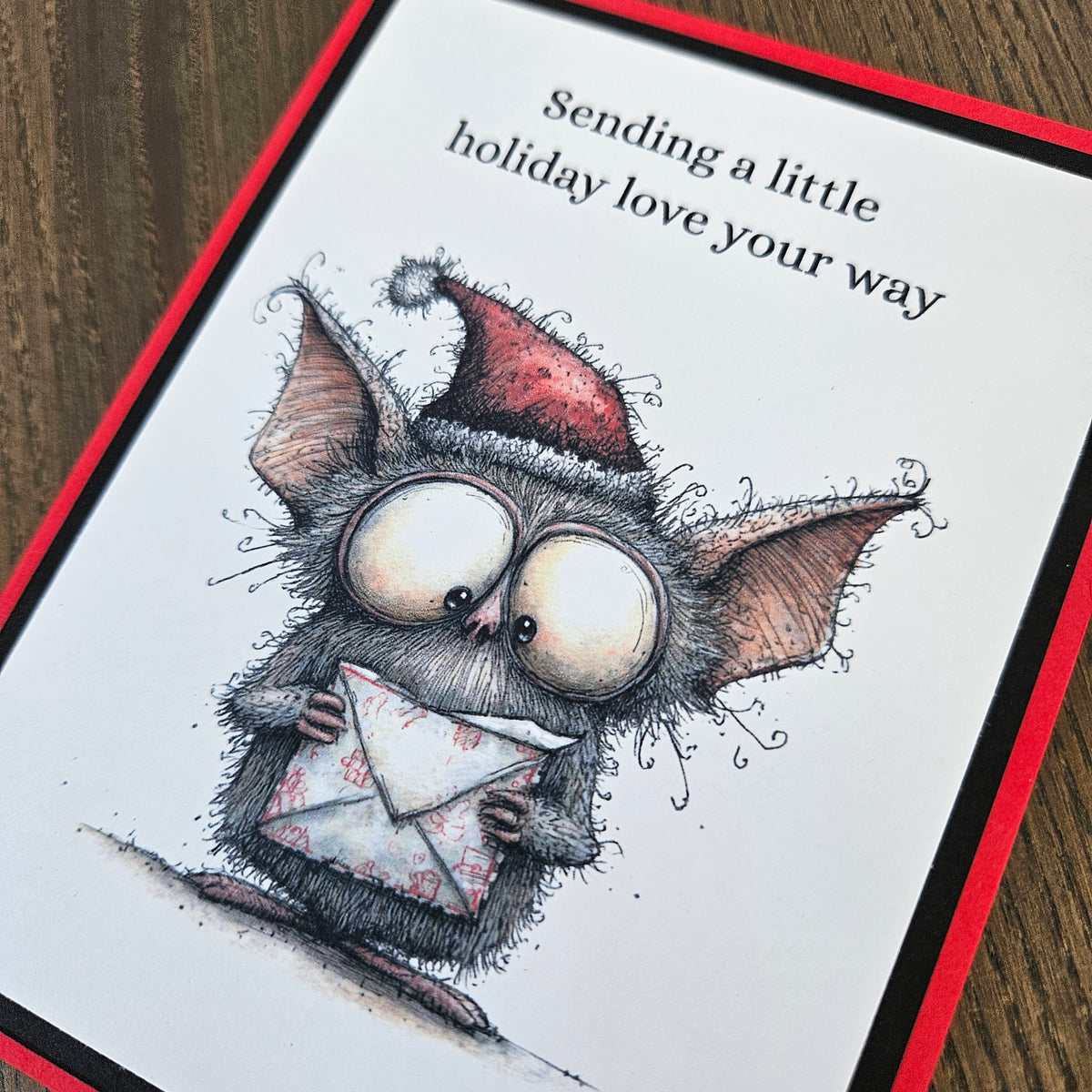 Angled close-up of wide-eyed bat in a Santa hat holding a patterned envelope