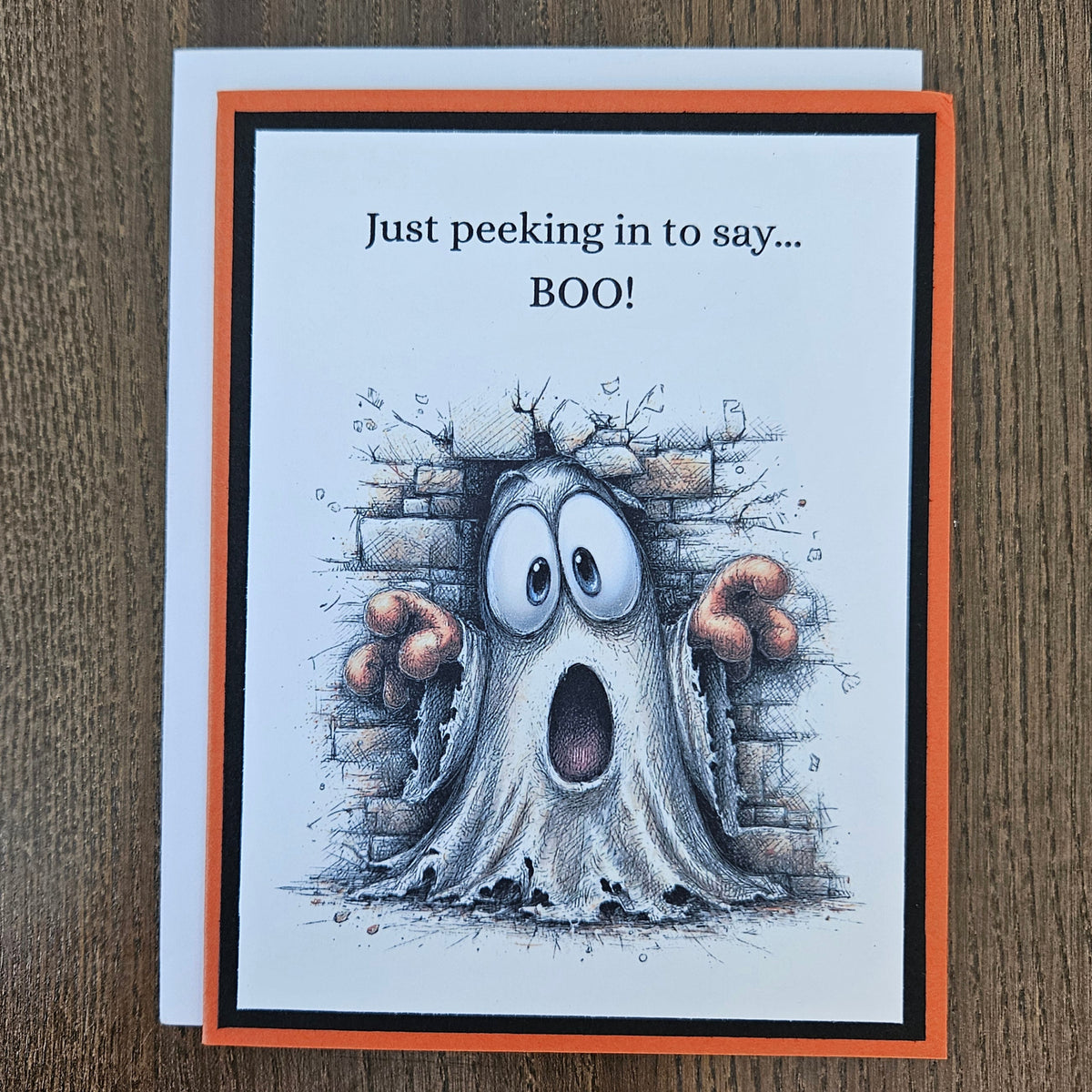 A square greeting card with a white front and orange border, placed on a brown wood surface. The card shows a cartoon ghost peeking out from behind a brick wall with the text 'Just peeking in to say.