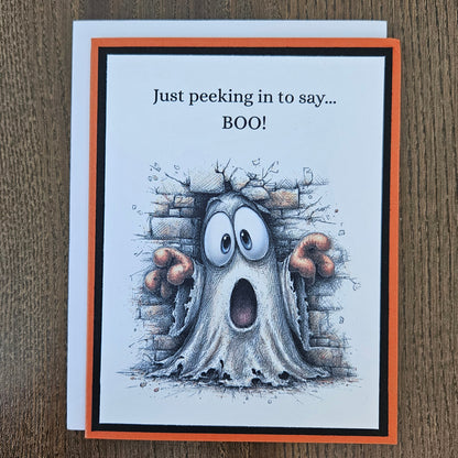 A square greeting card with a white front and orange border, placed on a brown wood surface. The card shows a cartoon ghost peeking out from behind a brick wall with the text 'Just peeking in to say.