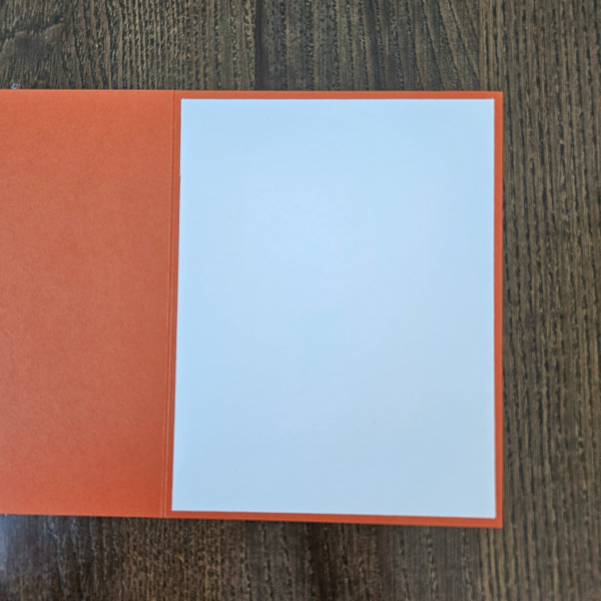 An open greeting card with a textured orange cover and a blank white insert, placed on a dark wood grain surface.