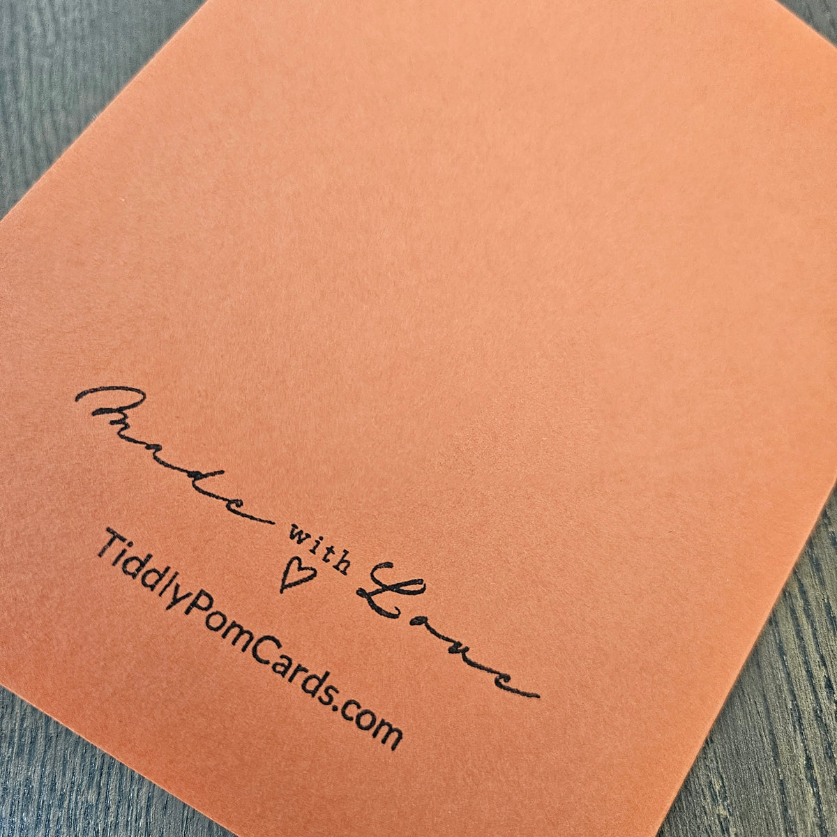 A close-up of a peach-colored pom pom card with black printed text reading 'Made with Love' and 'TiddlyPomCards.com' on a dark wood surface.