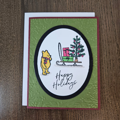 A handmade holiday greeting card featuring Winnie the Pooh on a sled with a Christmas tree and presents, set on a textured olive green background with a white envelope, photographed on a wooden surface.