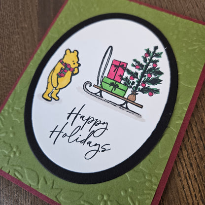 A handmade holiday greeting card featuring Winnie the Pooh in a yellow suit and scarf standing next to a sleigh carrying presents and a small Christmas tree, with the words 'Happy Holidays' in black script. The card has a white oval center, a black oval border, a textured olive green background, and a dark red outer border, photographed on a wooden surface.