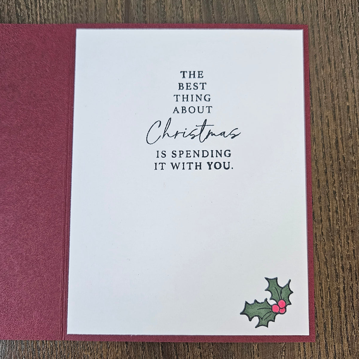 A square white Christmas greeting card with a burgundy border, placed on a wooden surface next to a matching burgundy envelope. The card features the printed message, 'THE BEST THING ABOUT Christmas IS SPENDING IT WITH YOU.