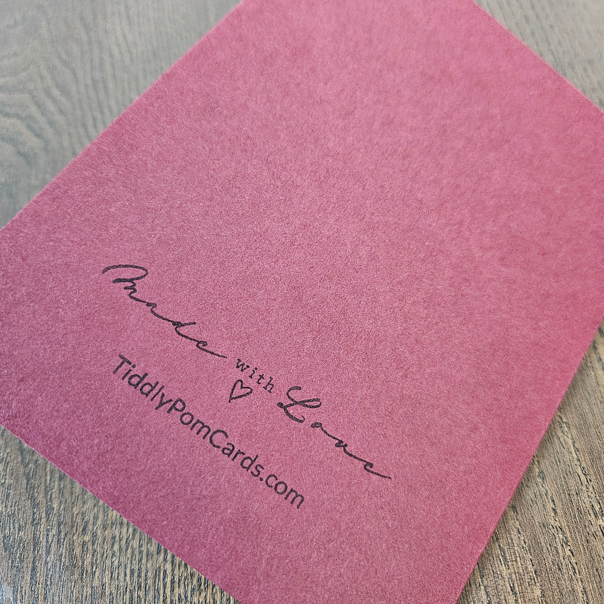 A square pink textured card or envelope with the words 'Made with Love' and the website 'TiddlyPomCards.com' printed in black script, shown on a gray wood-grain surface.