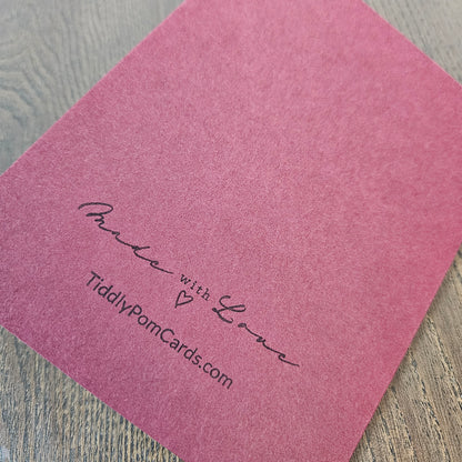 A square pink textured card or envelope with the words 'Made with Love' and the website 'TiddlyPomCards.com' printed in black script, shown on a gray wood-grain surface.